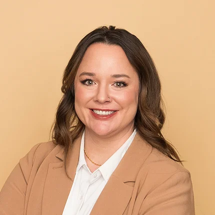 Woman with long brown hair wearing a tan blazer and white shirt, smiling in front of a plain light beige background.