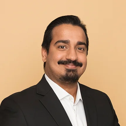 A man with dark hair and a mustache, wearing a black suit jacket and white shirt, smiles at the camera against a plain beige background.