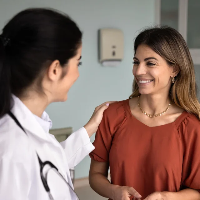 A doctor in a white coat with a stethoscope gently touches the shoulder of a smiling woman in a rust-colored top, as they have a friendly conversation in a medical office.