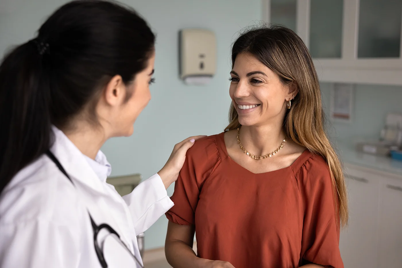 A female doctor wearing a white coat gently touches the shoulder of a smiling woman in an orange blouse as they talk in a medical office.