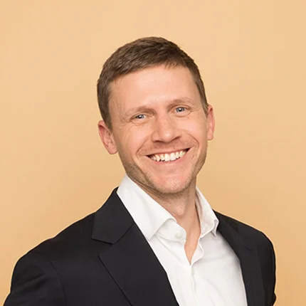 A smiling man with short light brown hair wearing a white dress shirt and a dark blazer, posing in front of a plain beige background.