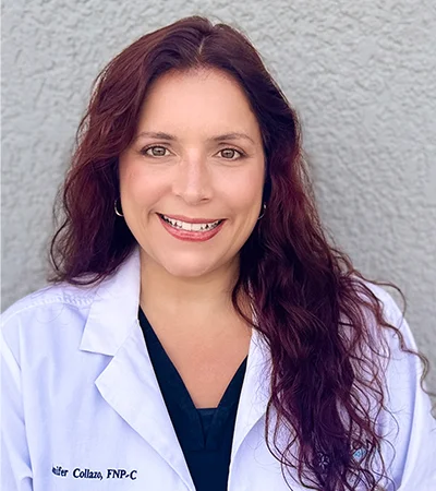 A woman with long, wavy brown hair smiles at the camera. She is wearing a white medical coat over dark scrubs and standing in front of a textured gray wall.