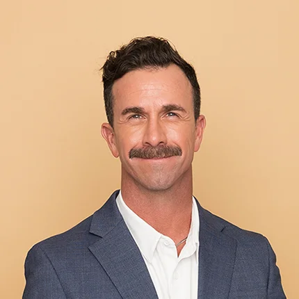 A man with short dark hair and a mustache is smiling at the camera. He is wearing a blue suit jacket over a white collared shirt, standing in front of a plain beige background.