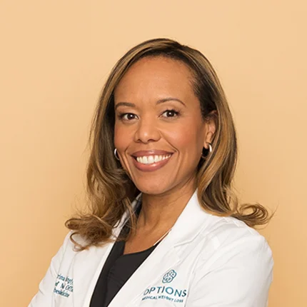 A smiling woman with long, light brown hair wearing a white medical coat stands against a plain beige background, arms crossed confidently.