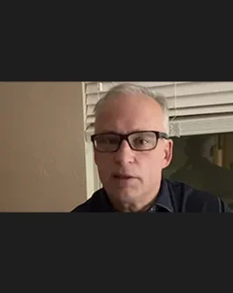 A middle-aged man with short gray hair and black glasses is indoors, standing in front of a window with closed blinds. He is wearing a dark shirt and looking directly at the camera.