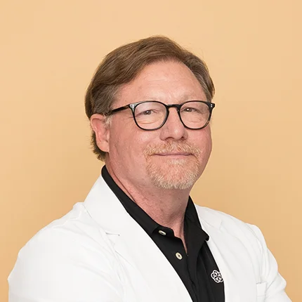 A middle-aged man with light brown hair, glasses, and a mustache and beard smiles at the camera. He is wearing a white coat over a black collared shirt, standing against a plain beige background.