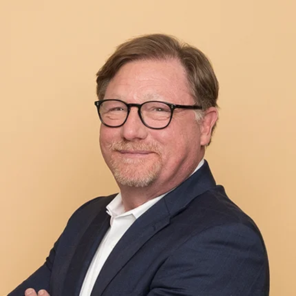A man with light brown hair and glasses, wearing a navy suit and white shirt, stands with arms crossed and smiles in front of a plain beige background.