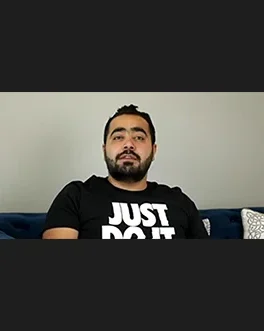 A man with dark hair and a beard sits on a blue couch, wearing a black t-shirt with the words JUST DO IT printed in white. The background is a plain light-colored wall with a patterned pillow visible.