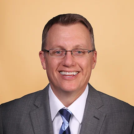 A man with short brown hair and glasses, wearing a gray suit, white shirt, and blue striped tie, smiles at the camera against a plain beige background.