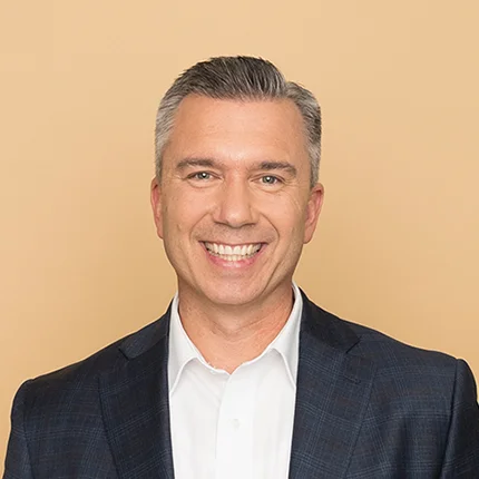 A middle-aged man with short gray hair, wearing a dark suit jacket and white dress shirt, smiling in front of a plain beige background.