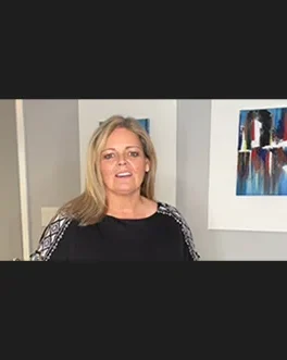 A woman with shoulder-length blonde hair, wearing a black top with patterned sleeves, stands indoors in front of a gray wall with two abstract art pieces hanging behind her.