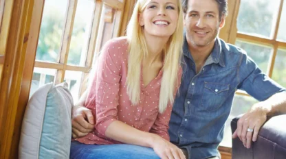 A smiling couple sits together on a couch by large windows. The woman has long blonde hair and wears a pink polka dot shirt, while the man has short dark hair and wears a denim shirt. They look happy and relaxed.