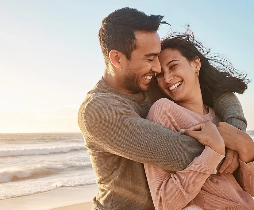 A couple stands on a beach at sunset, embracing and laughing together. The ocean waves and a clear sky are in the background, creating a warm, joyful atmosphere.