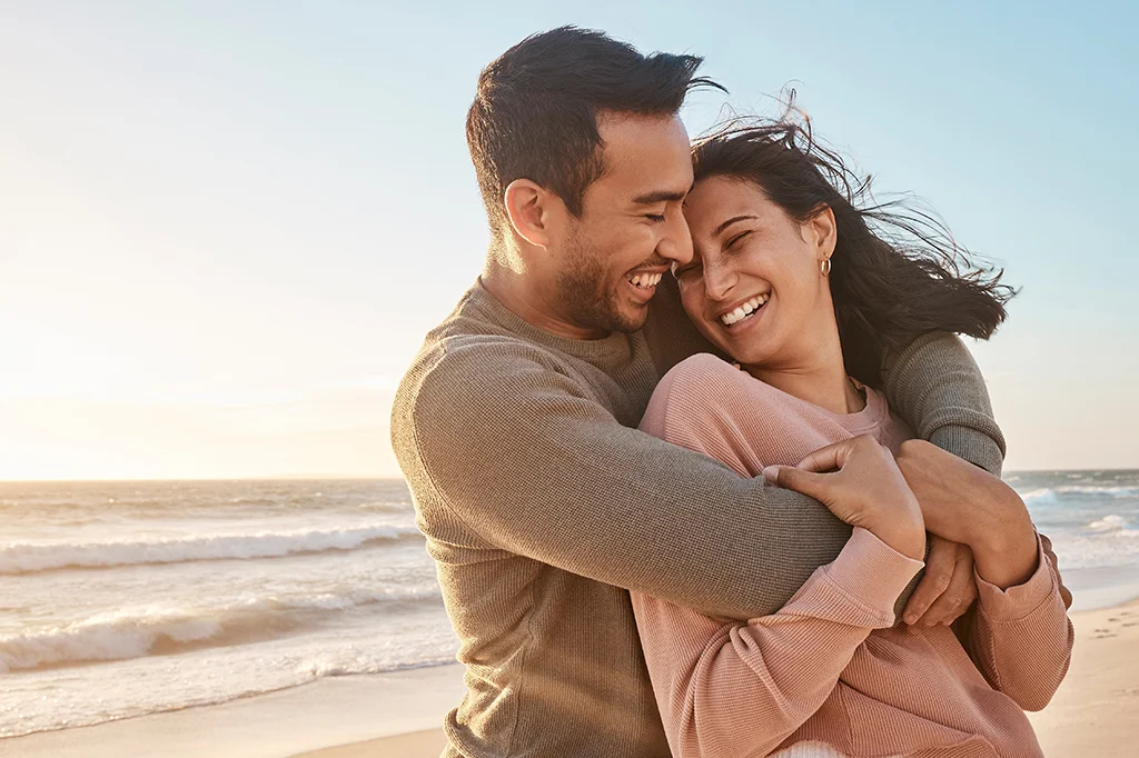 A couple stands on a beach at sunset, embracing and laughing together. The ocean waves and a clear sky are in the background, creating a warm, joyful atmosphere.