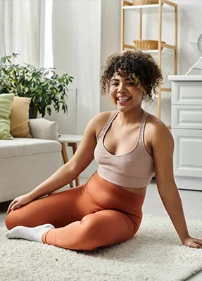 A smiling woman in a sports bra and orange leggings sits cross-legged on a rug in a bright living room, with plants and furniture in the background.