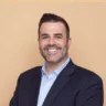 A man with short dark hair and a trimmed beard, wearing a dark blazer over a light blue shirt, smiles at the camera in front of a plain beige background.