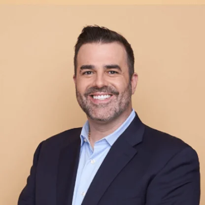 A man with short dark hair and a trimmed beard, wearing a dark blazer over a light blue shirt, smiles at the camera in front of a plain beige background.