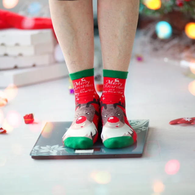 A person wearing festive Christmas socks stands on a digital scale near holiday decorations, including ornaments and gift boxes, with colorful lights in the foreground.