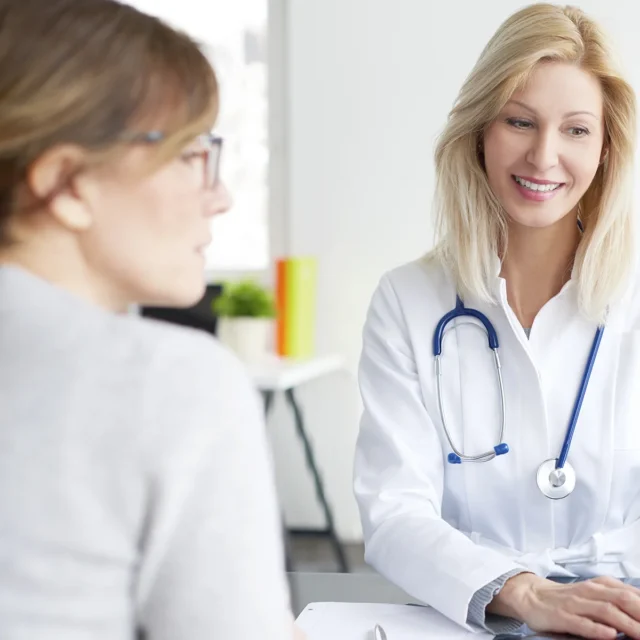 A smiling female doctor with a stethoscope talks to a patient across a desk in a bright, modern office setting.