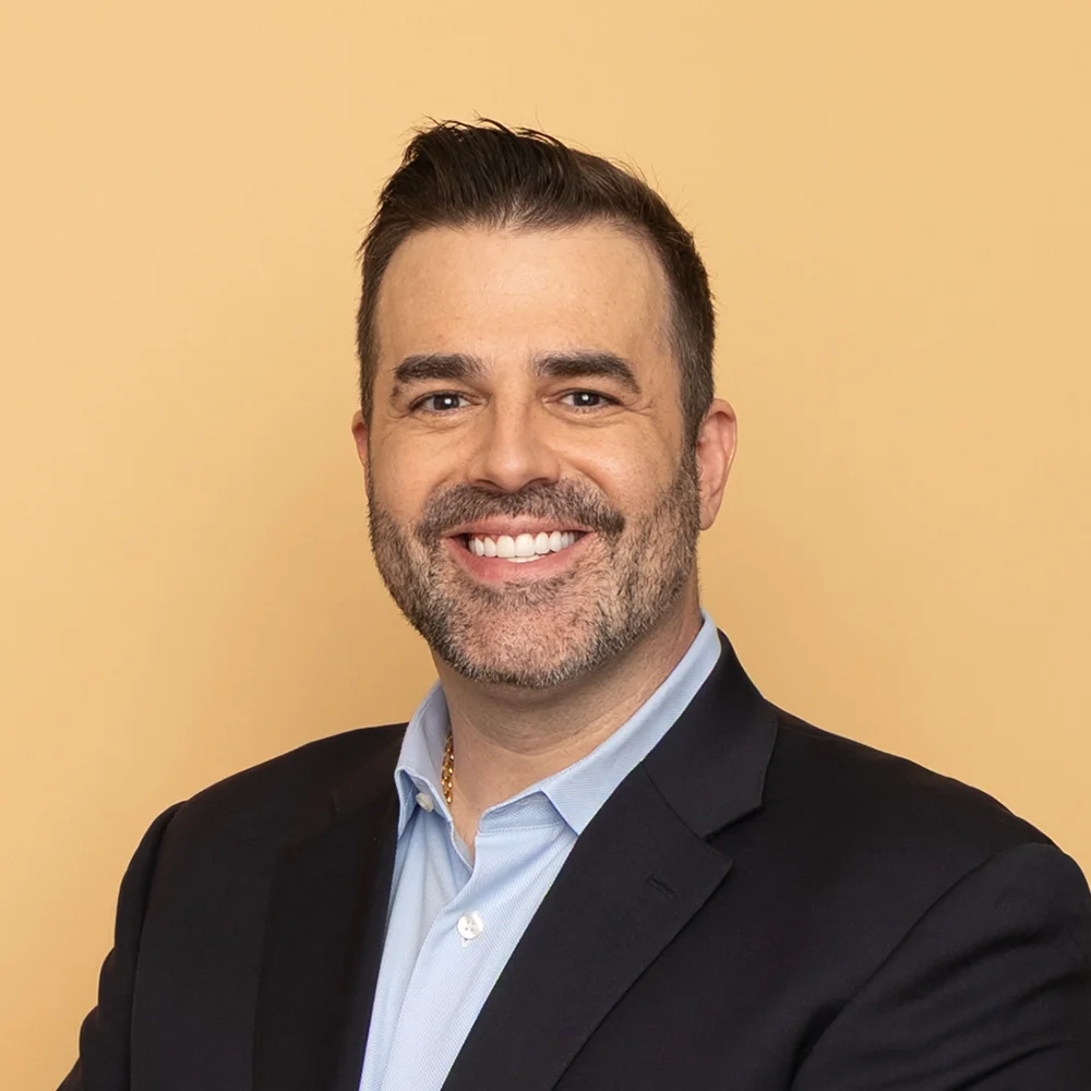 A man with short dark hair and a trimmed beard smiles at the camera. He is wearing a dark suit jacket over a light blue collared shirt, standing against a plain light tan background.