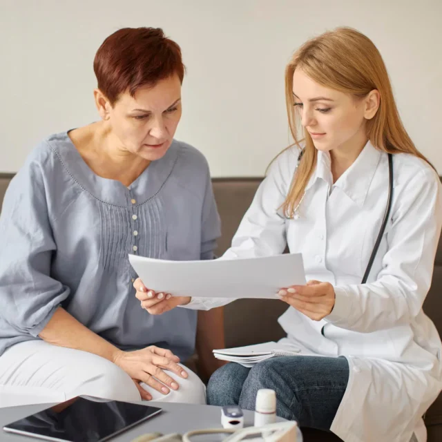 A female doctor shows and discusses medical documents with a seated middle-aged woman, both looking focused. Medical tools and a tablet are visible on the table in front of them.