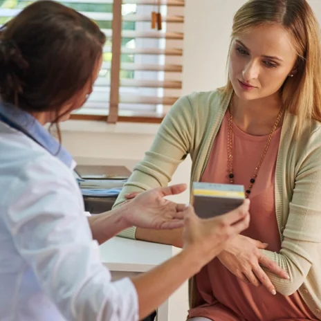 A doctor explains something to a concerned woman during a consultation in a well-lit office. The doctor is holding a small container or device, while the woman listens attentively with her arms folded.