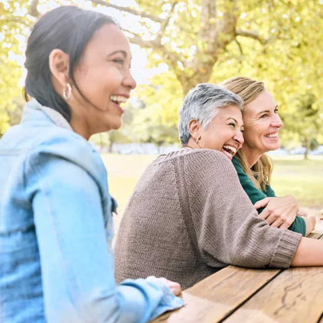 Three women sit at a wooden picnic table outdoors, smiling and laughing together on a sunny day, surrounded by green trees.