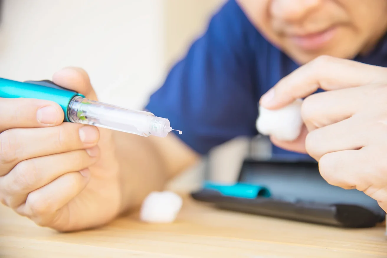 A person prepares an insulin pen for injection, holding the pen with a droplet at the needle tip and another hand holding a cotton ball, with a blurred case on the table nearby.