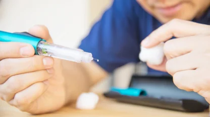 A person prepares an insulin pen for injection, holding the pen with a droplet at the needle tip and another hand holding a cotton ball, with a blurred case on the table nearby.