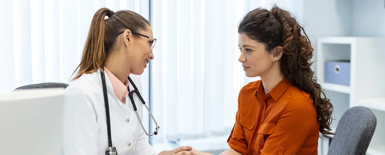 A doctor wearing a white coat and stethoscope sits across from a woman in an orange shirt, holding her hand and speaking supportively in a bright, modern office.
