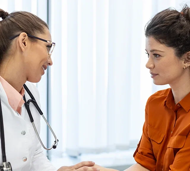 A doctor wearing a white coat and stethoscope sits across from a woman in an orange shirt, holding her hand and speaking supportively in a bright, modern office.