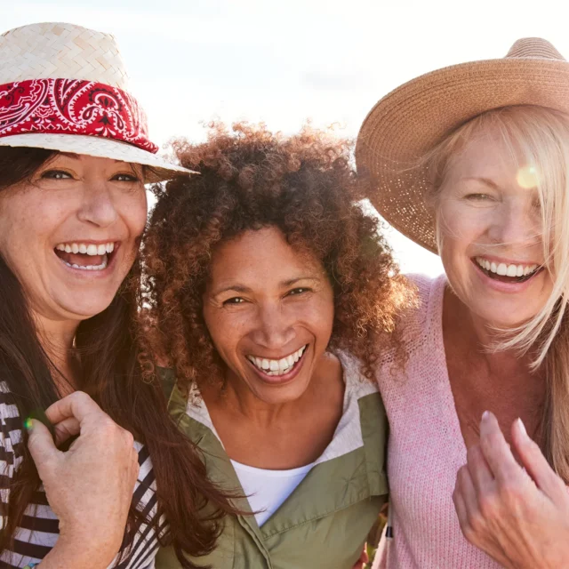 Three women outdoors smiling and laughing together. They are wearing casual clothes and hats, enjoying a sunny day, and their heads are close together in a friendly, joyful moment.