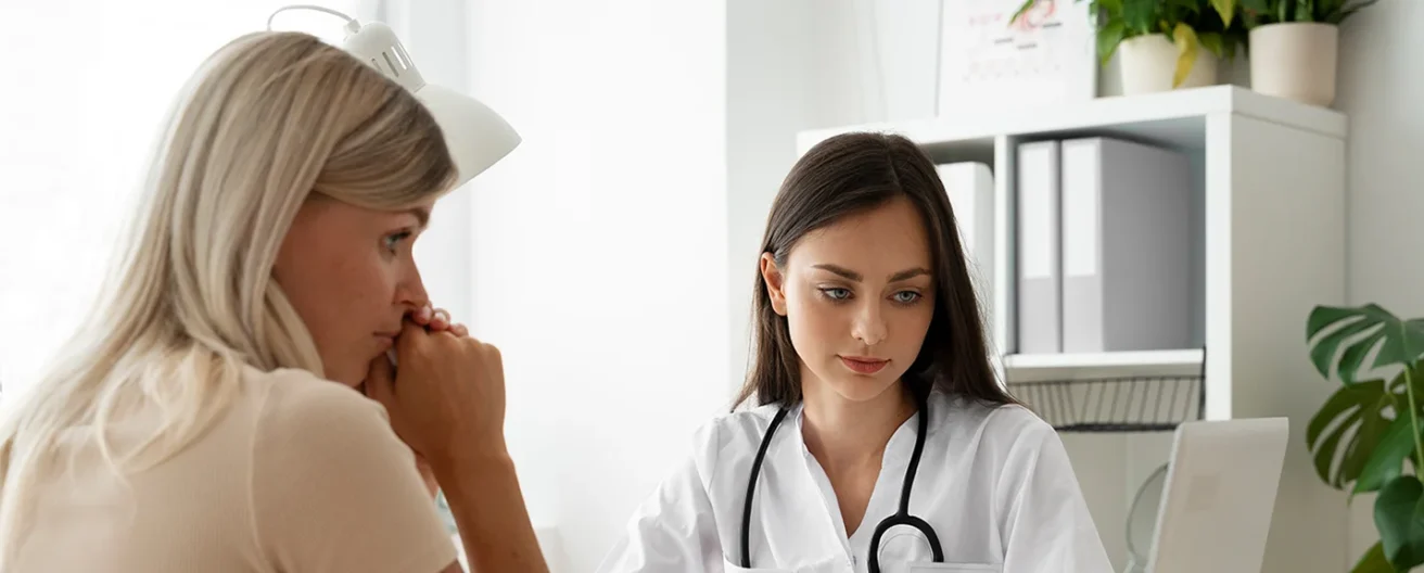 A doctor with a stethoscope talks to a concerned patient in a bright office, both women sitting at a desk with medical documents and a laptop nearby.