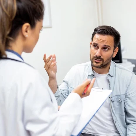 A male patient gestures while speaking to a female doctor who is holding a clipboard and pen, having a serious conversation in a medical office.