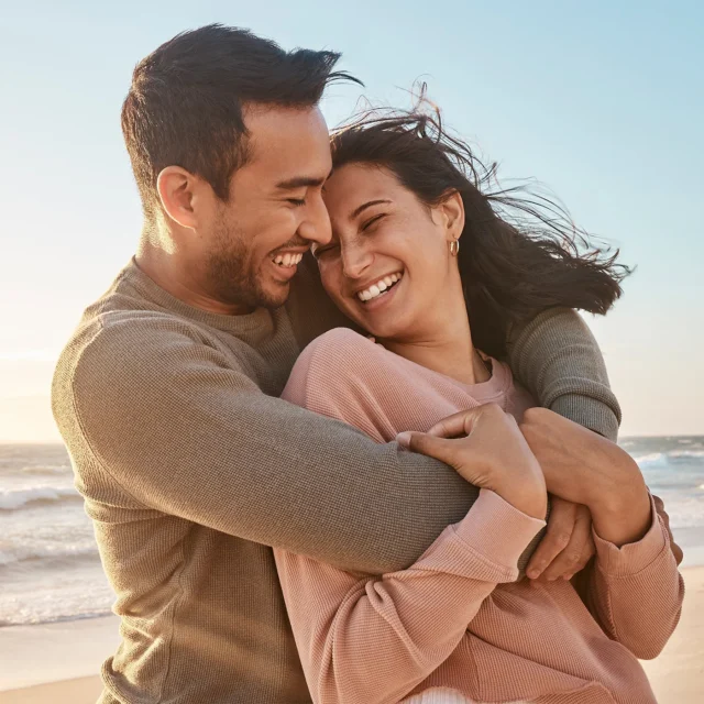 A couple stands on a sunny beach, smiling and embracing each other closely. The woman wears a pink sweater and the man wears a beige sweater. The ocean and sky are visible in the background.