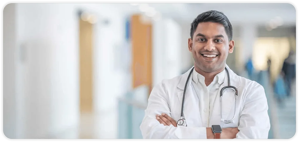 A smiling doctor with dark hair, wearing a white coat and stethoscope, stands with arms crossed in a bright hospital hallway.