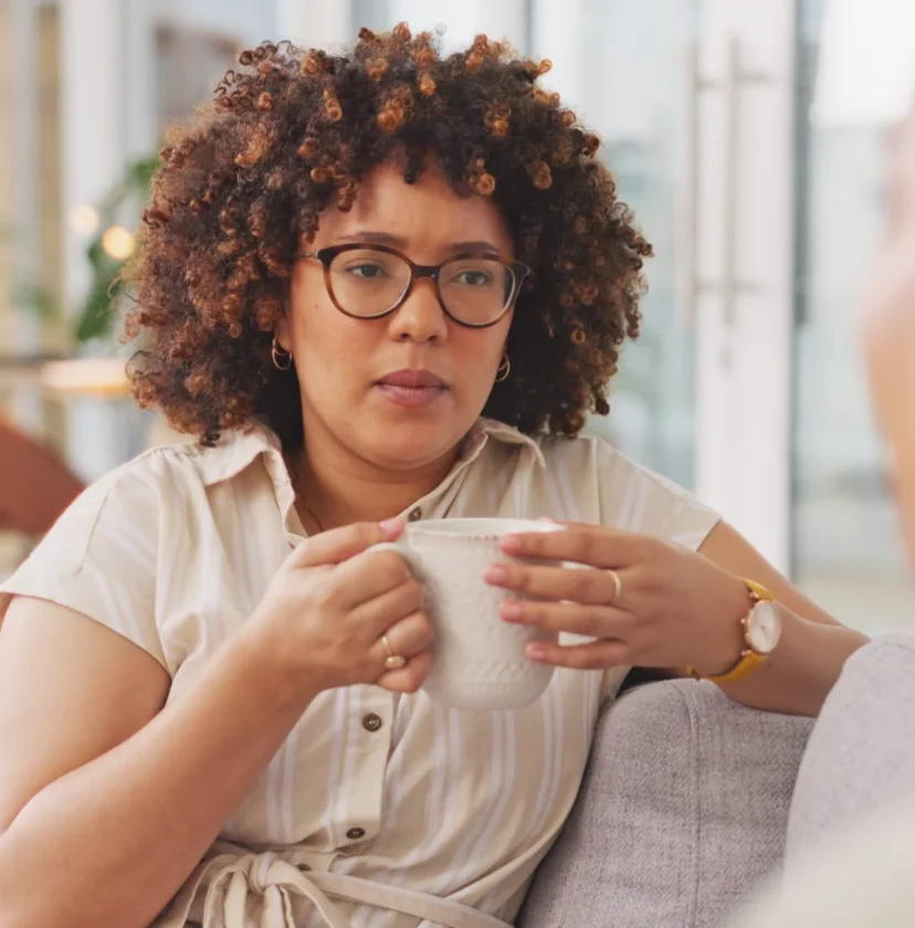 A woman with curly hair and glasses sits on a sofa holding a mug, listening attentively to another person who is partially visible in the foreground. The setting appears to be a relaxed, well-lit living room or office.