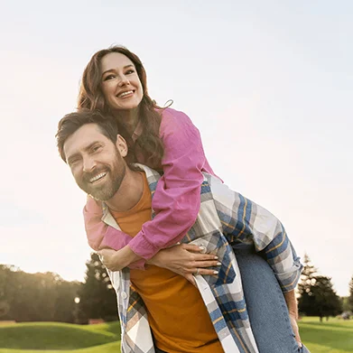 A smiling man gives a piggyback ride to a cheerful woman outdoors on a grassy field, with trees and a clear sky in the background. Both appear happy and are casually dressed.