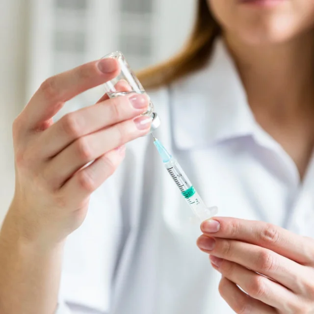 A healthcare professional fills a syringe with liquid from a glass vial, preparing for an injection. The person wears a white coat and the setting appears clinical.