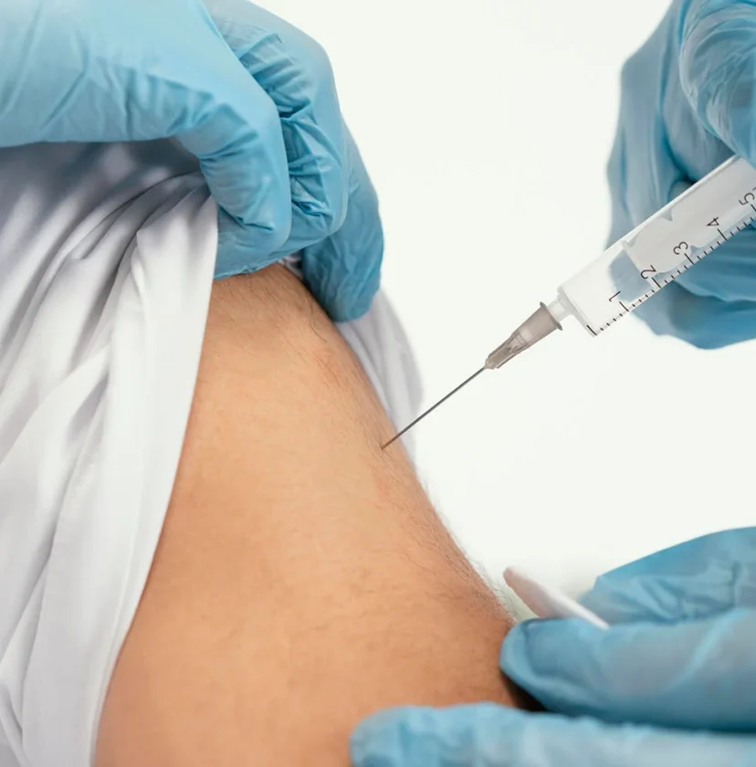 A close-up of a healthcare worker wearing blue gloves giving an injection with a syringe into a persons upper arm, who is pulling up their white sleeve.