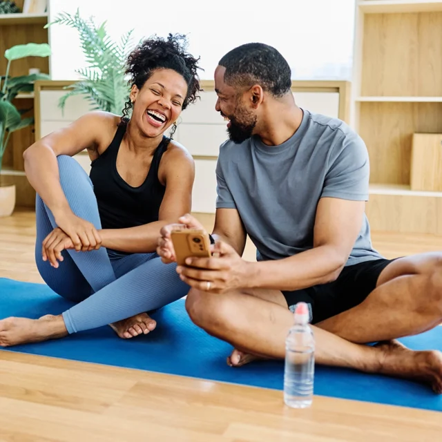 A smiling woman and man sit on a yoga mat indoors, laughing together. The man holds a smartphone, and a water bottle is in front of them. They appear relaxed and happy after exercising.