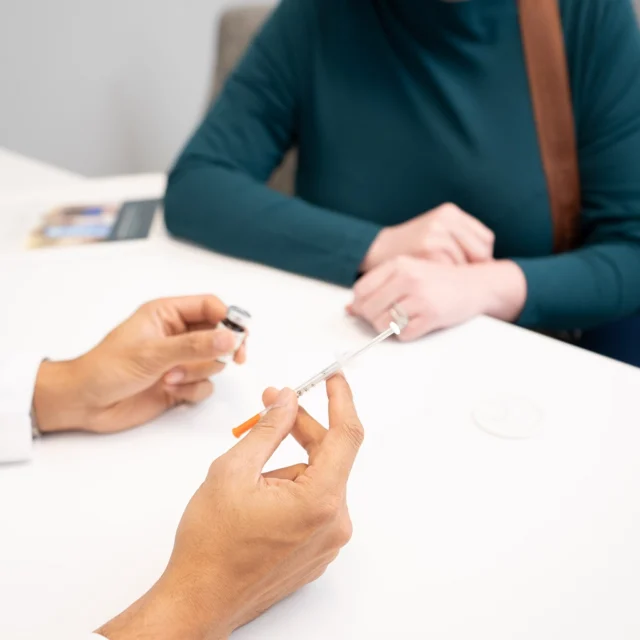 A healthcare professional holds a syringe and medication vial while speaking with a patient wearing a green top, seated at a table in a medical setting.