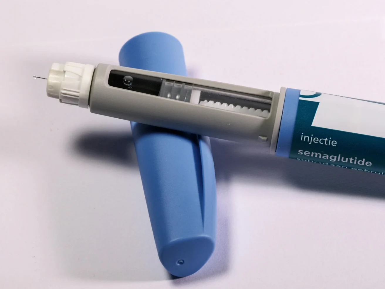A close-up of a semaglutide injection pen with its blue cap removed, showing the needle and dosage window, placed on a white surface.