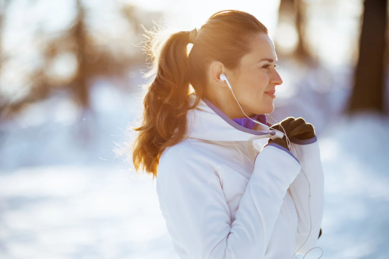 A woman in a white jacket and gloves stands outdoors in a snowy landscape, smiling and listening to earphones. The sun shines brightly behind her, creating a warm glow.