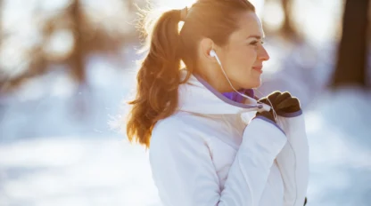 A woman in a white jacket and gloves stands outdoors in a snowy landscape, smiling and listening to earphones. The sun shines brightly behind her, creating a warm glow.