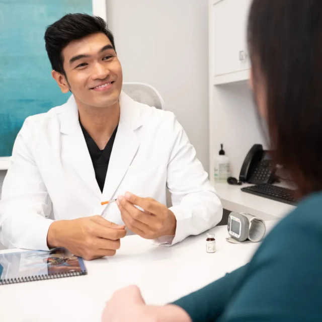 A doctor in a white coat smiles while discussing Medical Weight Loss options with a patient in an office. He holds a syringe, and medical supplies are on the desk between them. The patient is seated with their back to the camera.