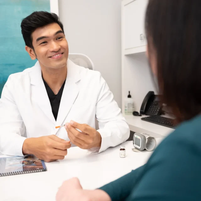 A male doctor in a white coat smiles while holding a syringe, speaking with a patient in an office. Medical supplies and a Medical Weight Loss brochure are on the desk between them.