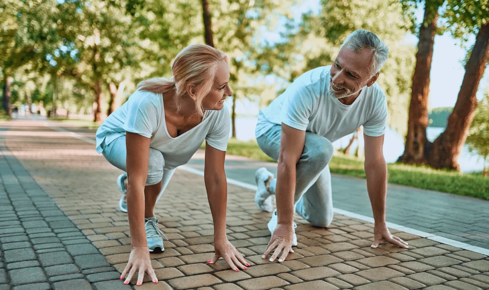 An older man and woman wearing athletic clothes smile at each other while in a starting position for a run on a paved path in a park with trees and sunlight in the background.