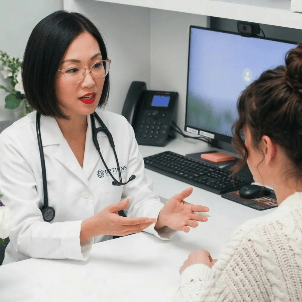 A female doctor wearing a white coat and stethoscope discusses medical weight loss with a patient across a desk in a medical office, with a computer, phone, and paperwork visible in the background.