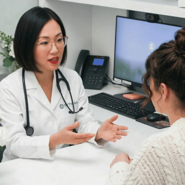 A female doctor wearing glasses and a white coat talks to a patient across a desk in a medical office, with a computer, phone, and notepad on the desk.